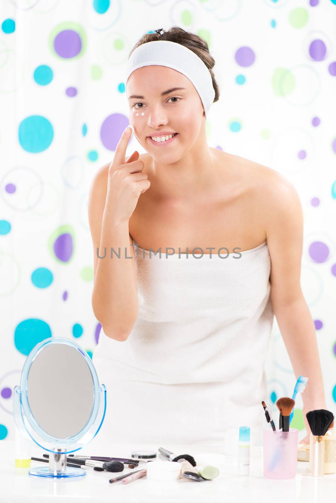 Young cute woman preparing to start her day. She is applying moisturizer cream on face. Looking at camera.