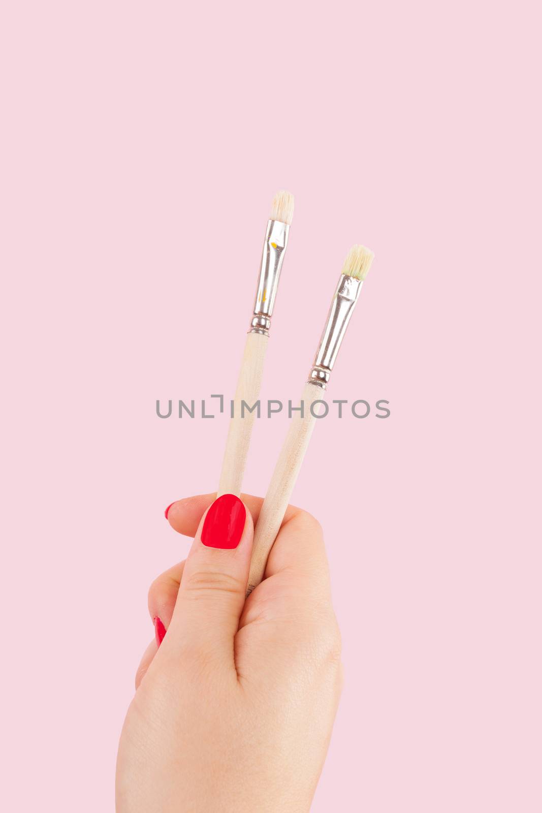 Female hand with red fingernails holding paintbrush isolated on pink background. Creative painting. 