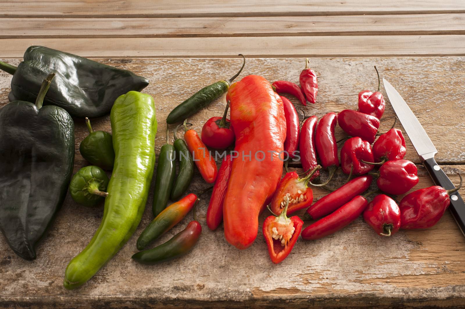 Variety of different chili peppers in a kitchen lying displayed by color on a wooden counter top with a knife waiting to be cut up for cooking