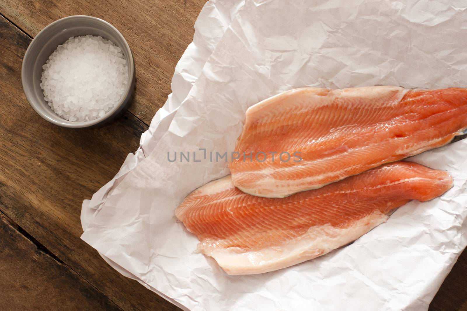 Two gourmet raw rainbow trout fillets lying on crumpled white paper with a ramekin of rock salt alongside waiting to be cooked, overhead view