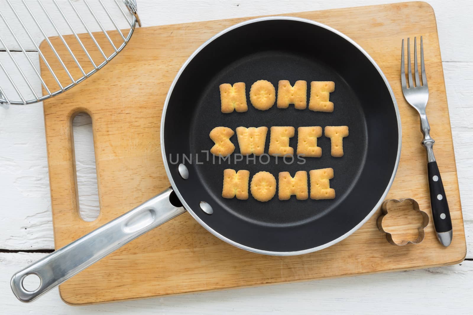 Top view of letter collage made of biscuits. Quote HOME SWEET HOME putting in black frying pan. Other cooking equipments: fork, cookie cutter and chopping board putting on white wooden table, vintage style image.