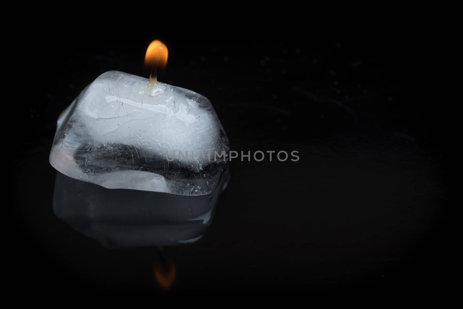 A close up shot of a lit candle wick stuck into an ice cube on a black background with reflection.