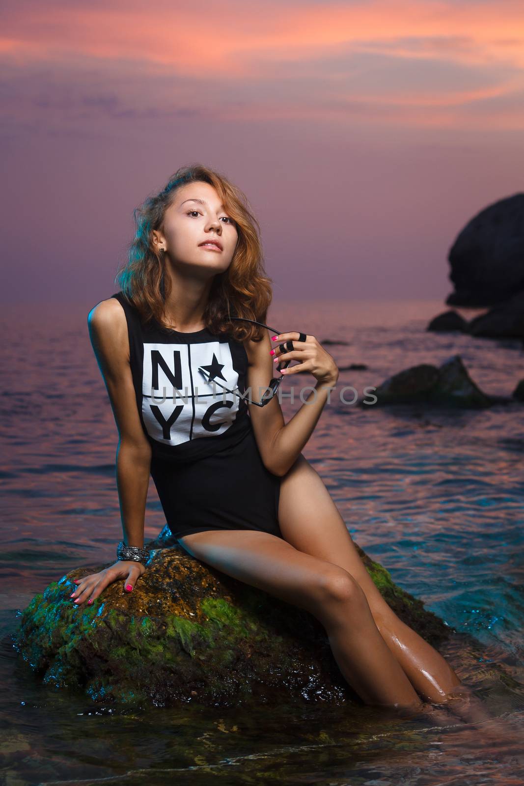 Beautiful young teen girl sitting on the stone in the sea fashion shoot at sunset beach