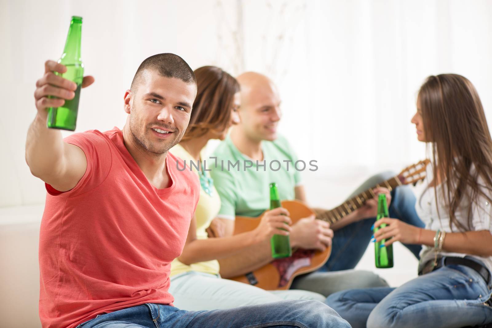 Close up of a young men smiling at home party with beer and cheers. Her friends in the background.