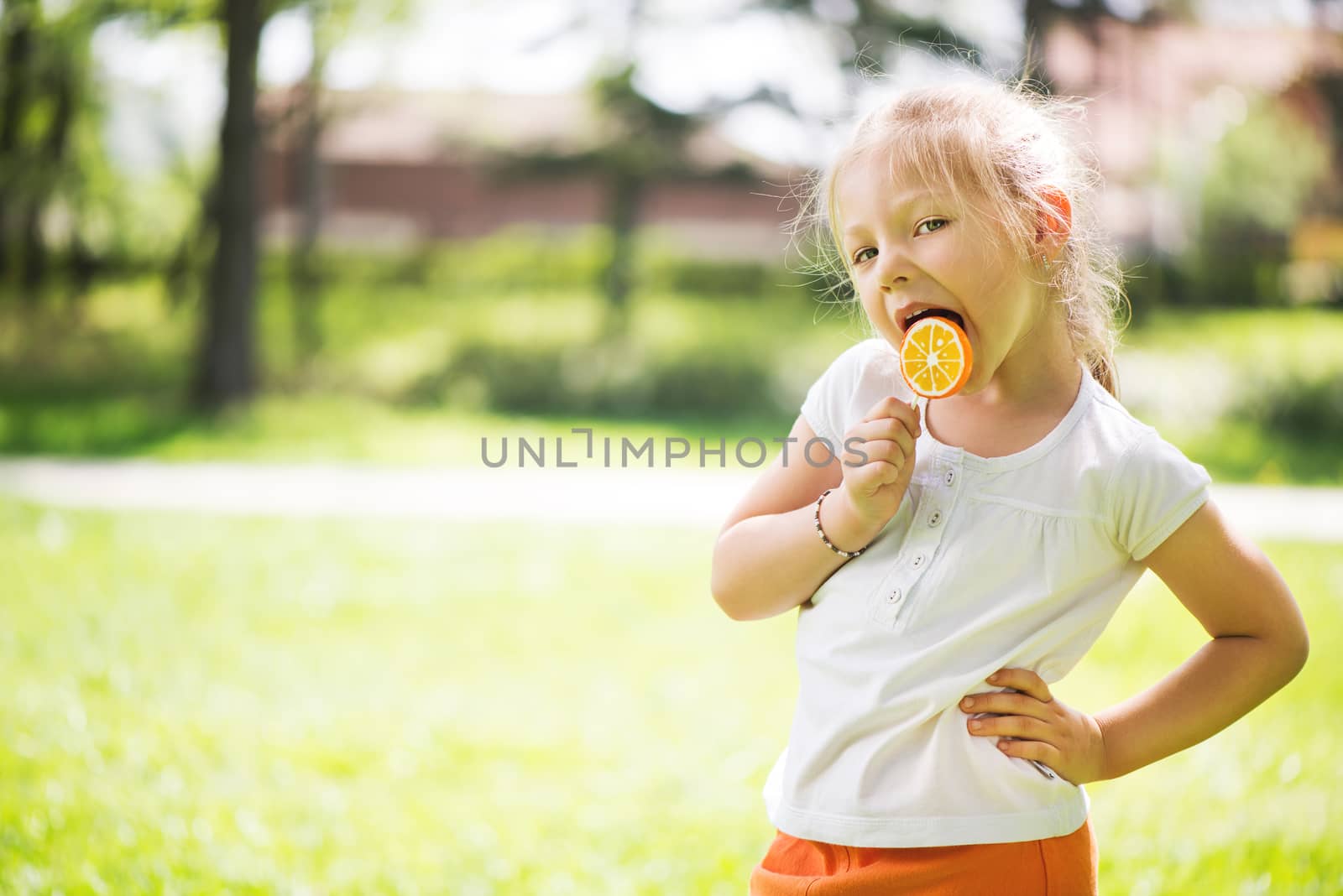 Cute Little Girl Standing on Green Grass in the Park and holding in hand orange Lollipop.