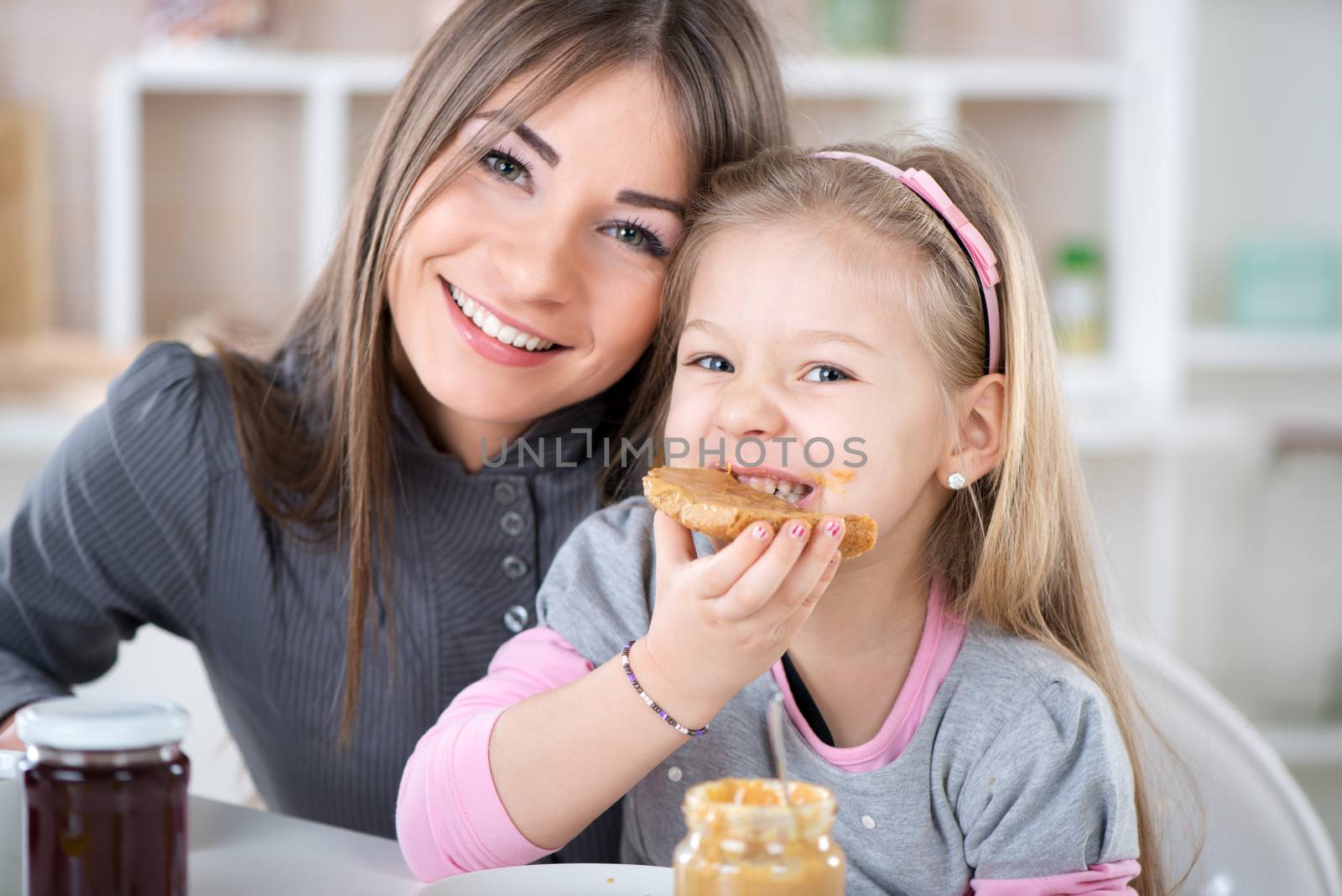 Mother and daughter breakfast in the kitchen. Cute little girl eats bread with peanut butter. Looking at Camera.