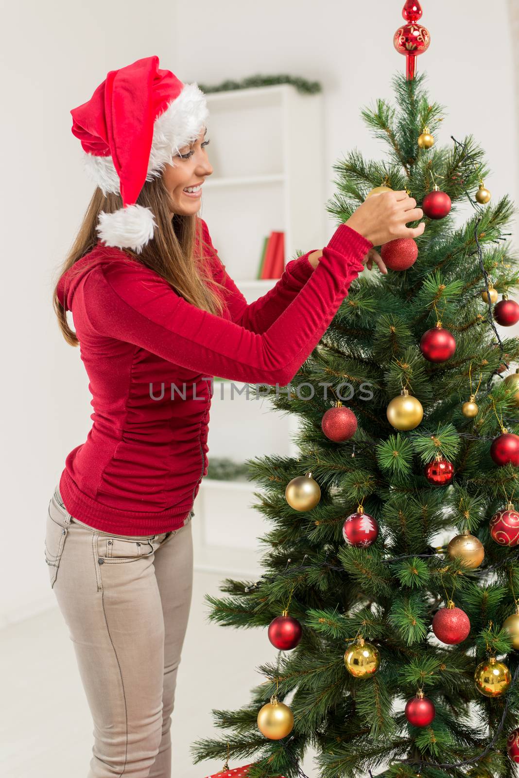 Happy beautiful young woman decorating christmas tree with red ornament at home.