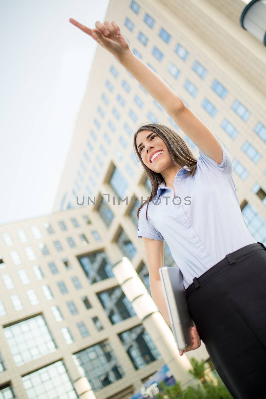 Young beautiful business woman with laptop in her hands, standing in front of office building, smile looking into the distance and finger pointing to the heights.