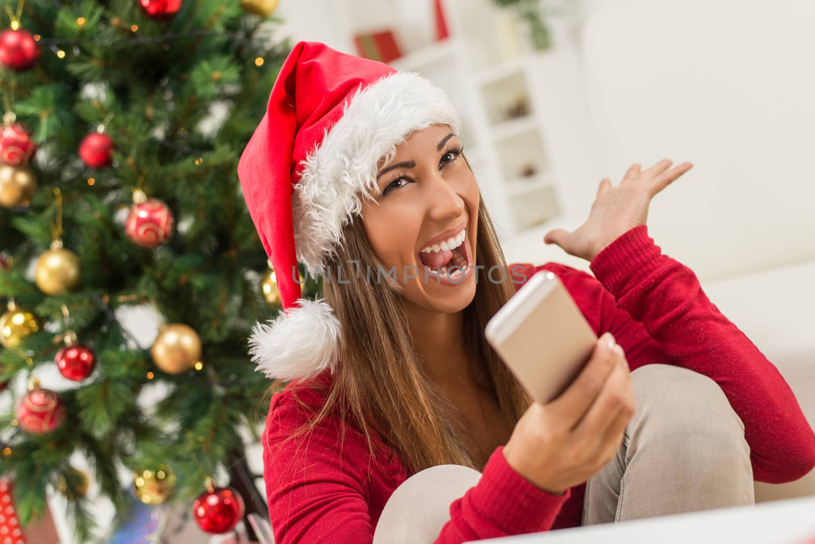 Cheerful young woman wearing Santa's hat is holding a smart phone at Christmas Time.