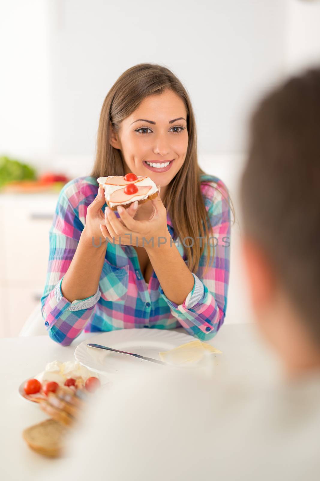 Beautiful young couple having breakfast in the domestic kitchen. Girlfriend eating sandwich and looking her boyfriend.