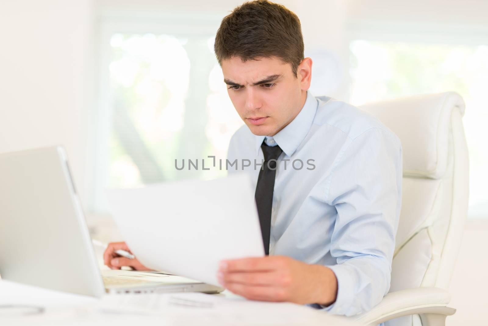 Young businessman sitting at an office desk in front of a laptop and looking at document.