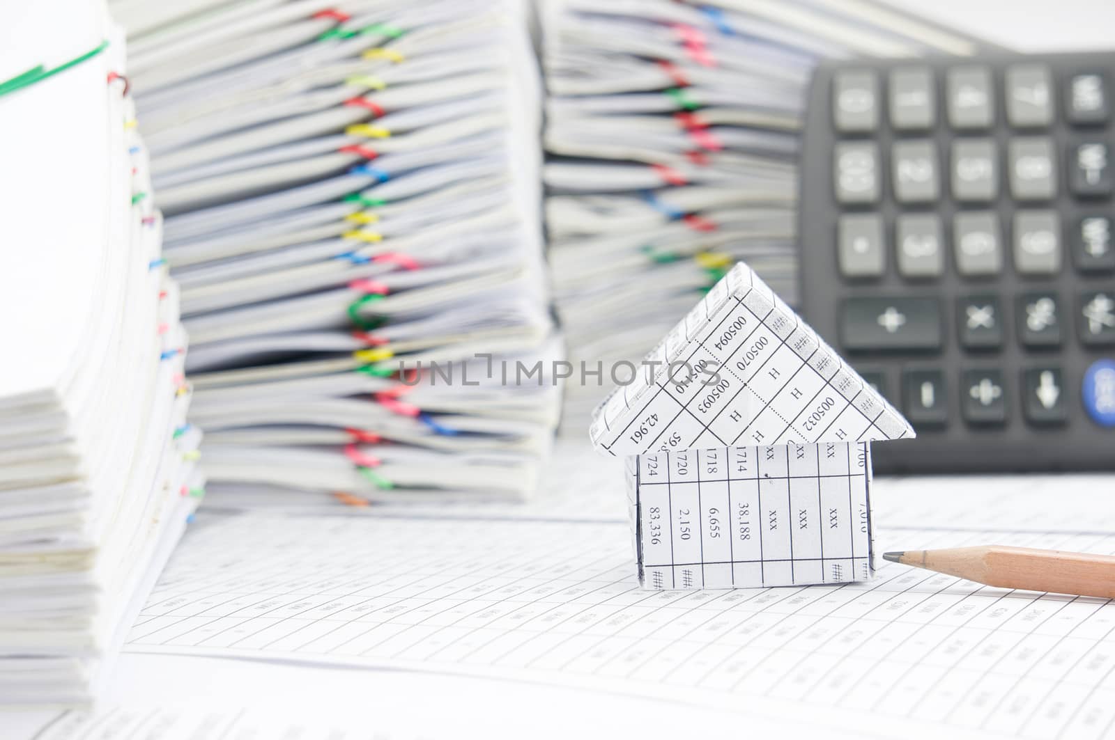 House and brown pencil on finance account have blur calculator and pile of paperwork as foreground and background.