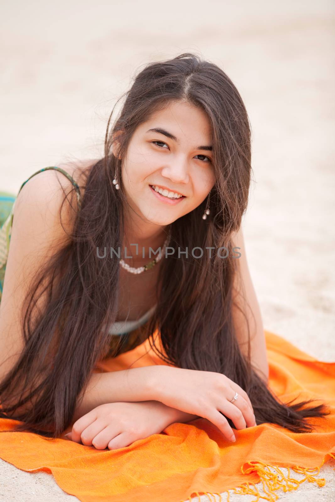 Beautiful biracial Asian Caucasian teen girl lying on orange blanket at beach, resting up on elbows 