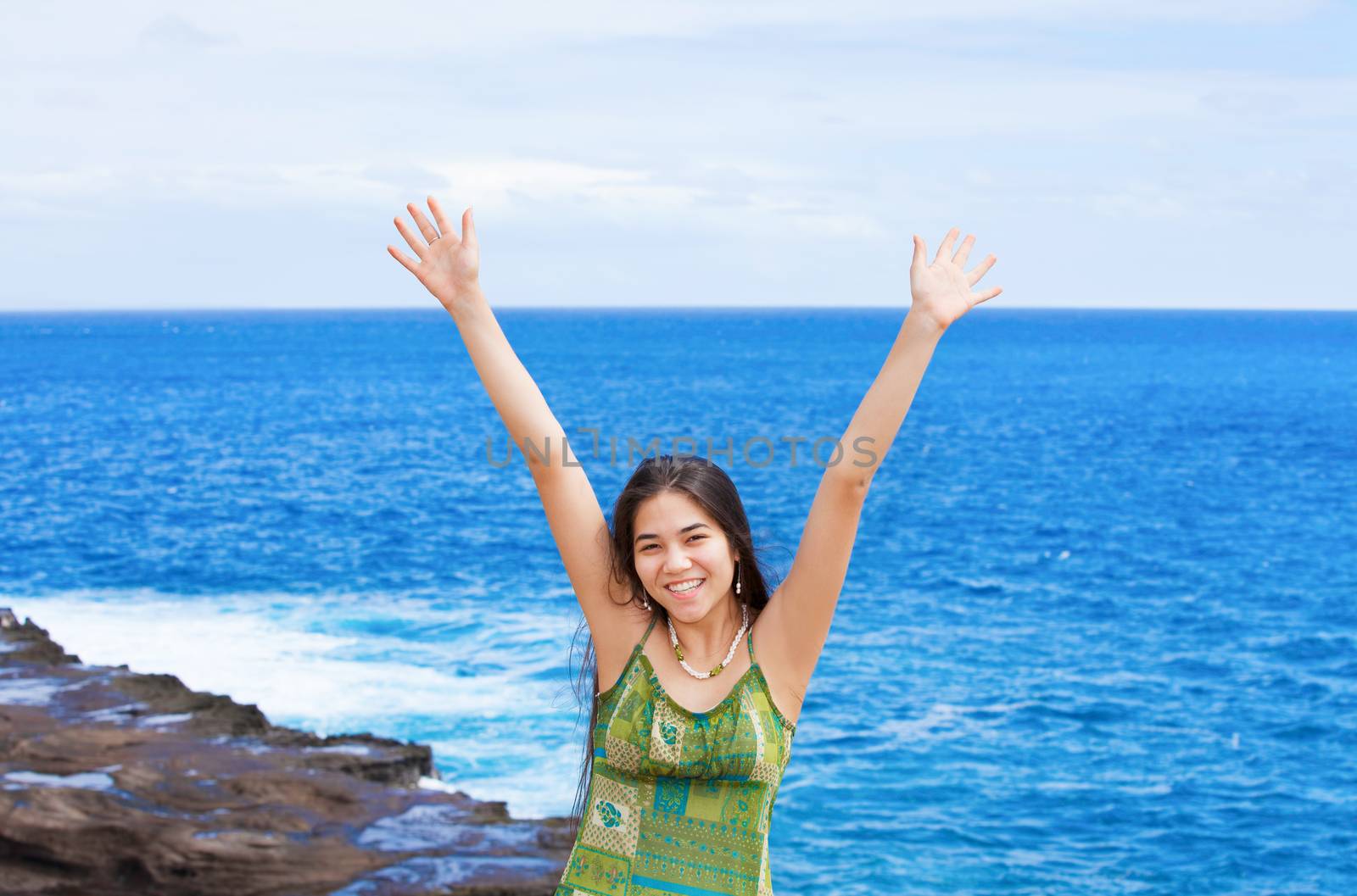 Beautiful biracial Asian Caucasian teen girl arms raised upwards in praise by blue ocean waters along rocky shore