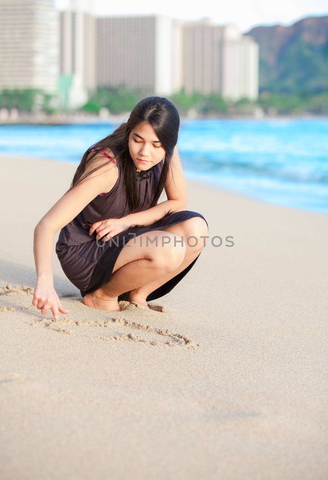 Beautiful biracial Asian Caucasian teen girl crouching on Waikiki beach, Hawaii, drawing with her finger in the sand as a wave comes onto shore. Urban scene and Diamond Head in background