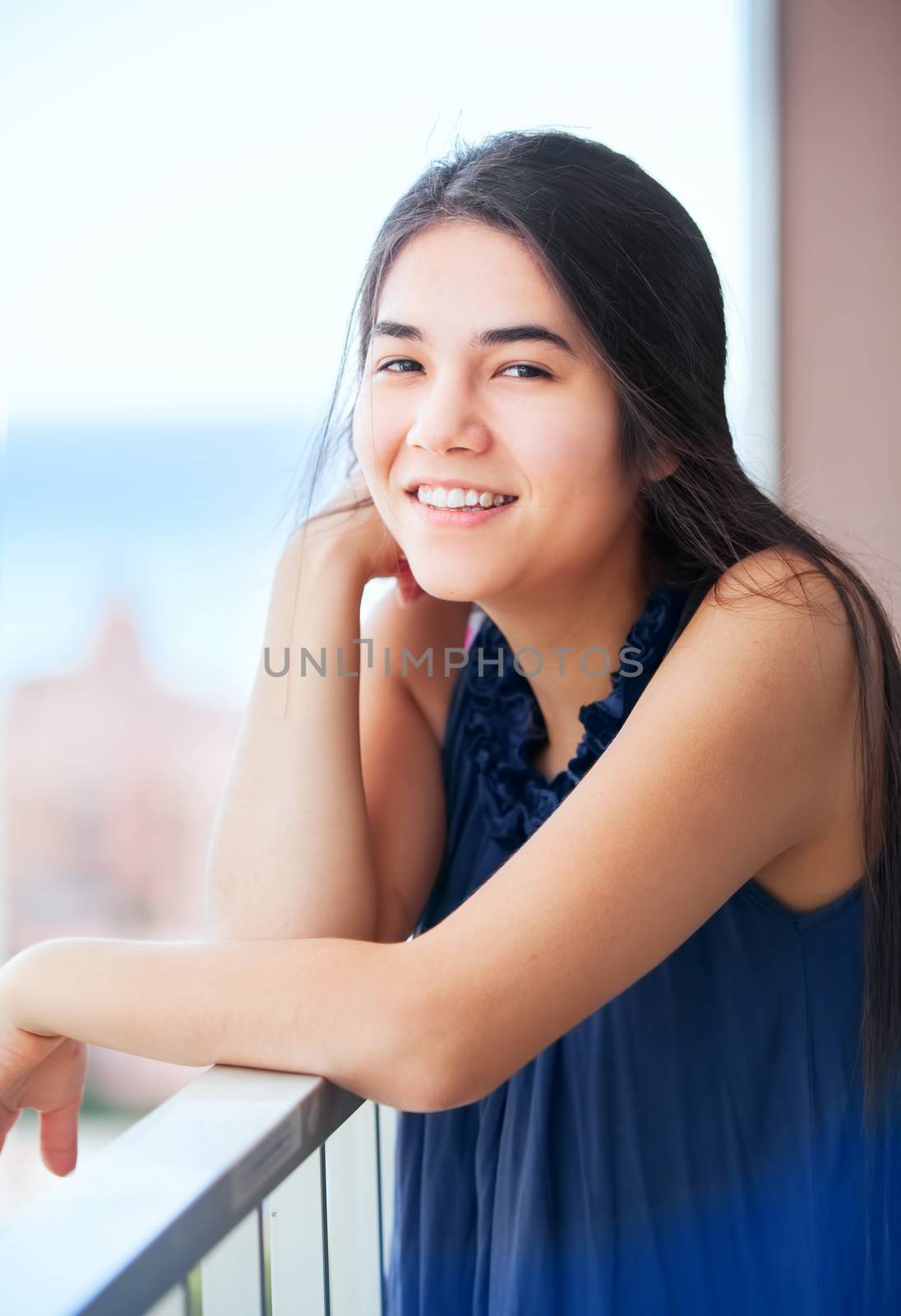 Beautiful biracial Asian Caucasian teen girl standing on outdoor high rise patio deck, leaning on railing with ocean landscape in background