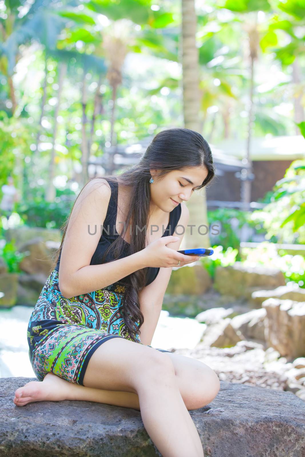 Beautiful biracial Asian Caucasian teen girl sitting on rock in tropical setting looking at cellphone, palm trees in background