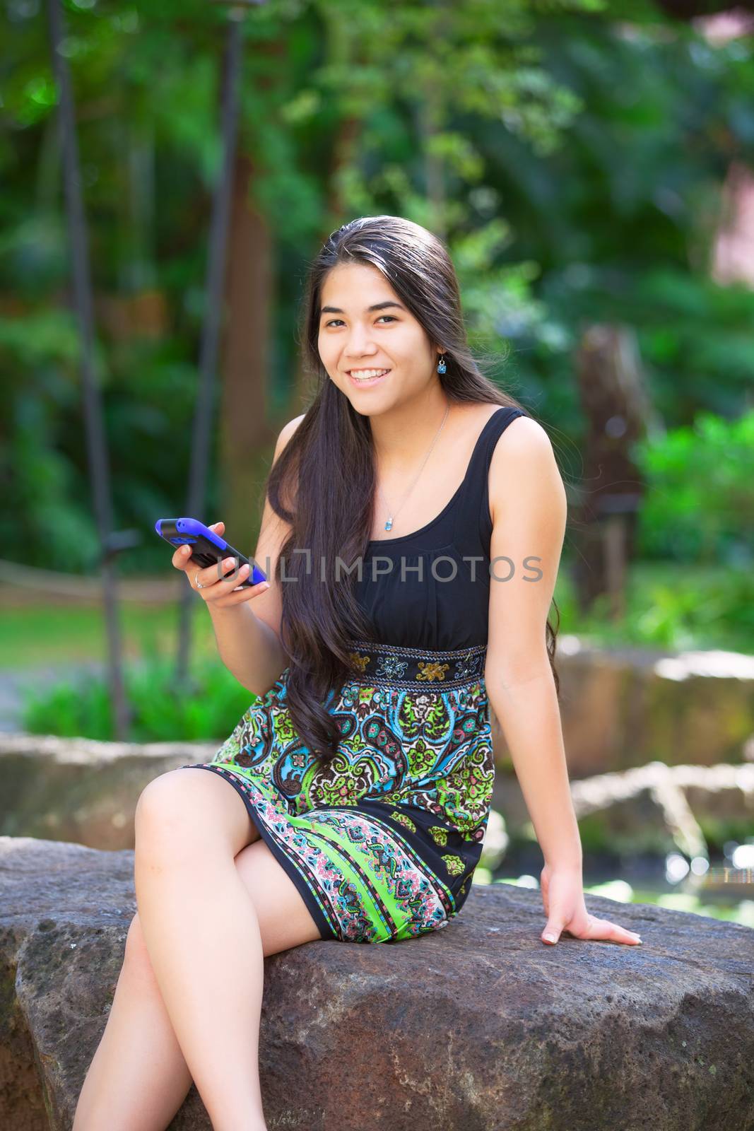 Beautiful biracial Asian Caucasian teen girl sitting on rock in tropical setting looking at cellphone, palm trees in background