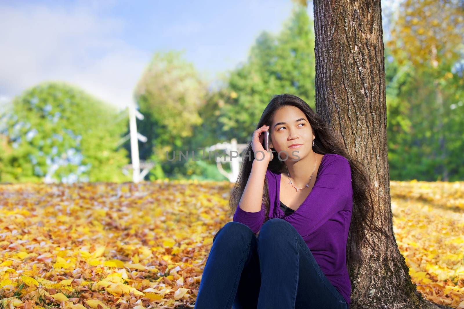 Teen girl sitting against autumn tree using cell phone by jarenwicklund