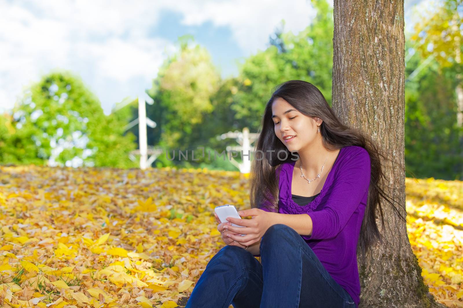 Teen girl sitting against autumn tree using cell phone by jarenwicklund