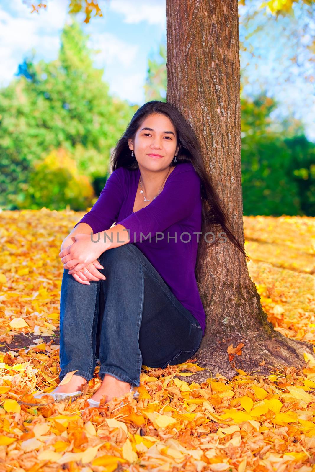 Beautiful biracial Asian Cuacasian teen girl sitting with back to tree, autumn leaves on ground
