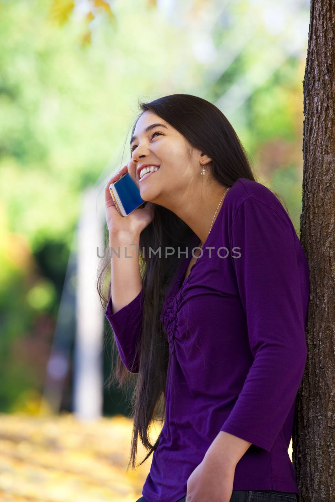 Beautiful biracial Asian Caucasian teen girl leaning against tree talking happily on cell phone, autumn leaves on ground