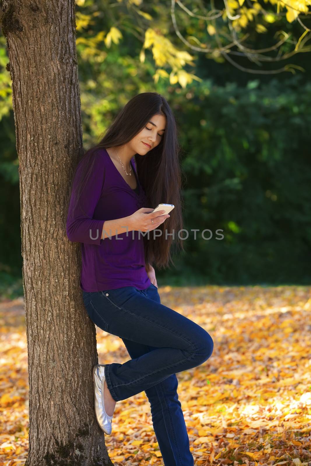 Teen girl standing against autumn tree looking at cell phone by jarenwicklund