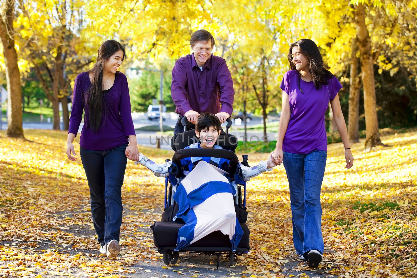 Multiracial family with disabled child in wheelchair walking among autumn leaves 