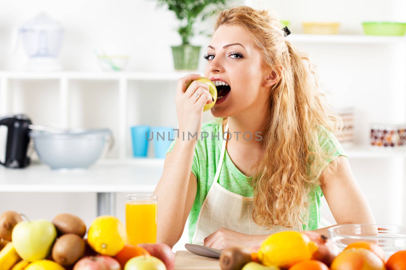 Beautiful young woman eating an apple in a kitchen. Looking at camera.