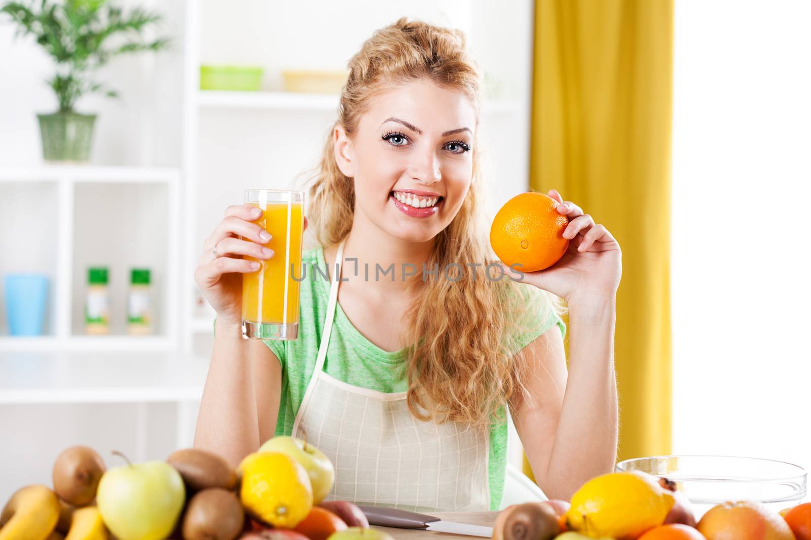 Beautiful young woman in a kitchen, holding an orange and juice. Looking at camera.