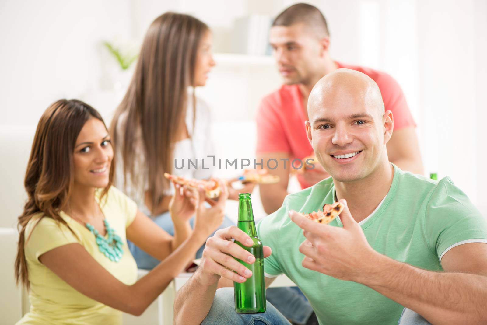 Close up of a young men smiling and eating pizza and drinking bear with her friends in the background.