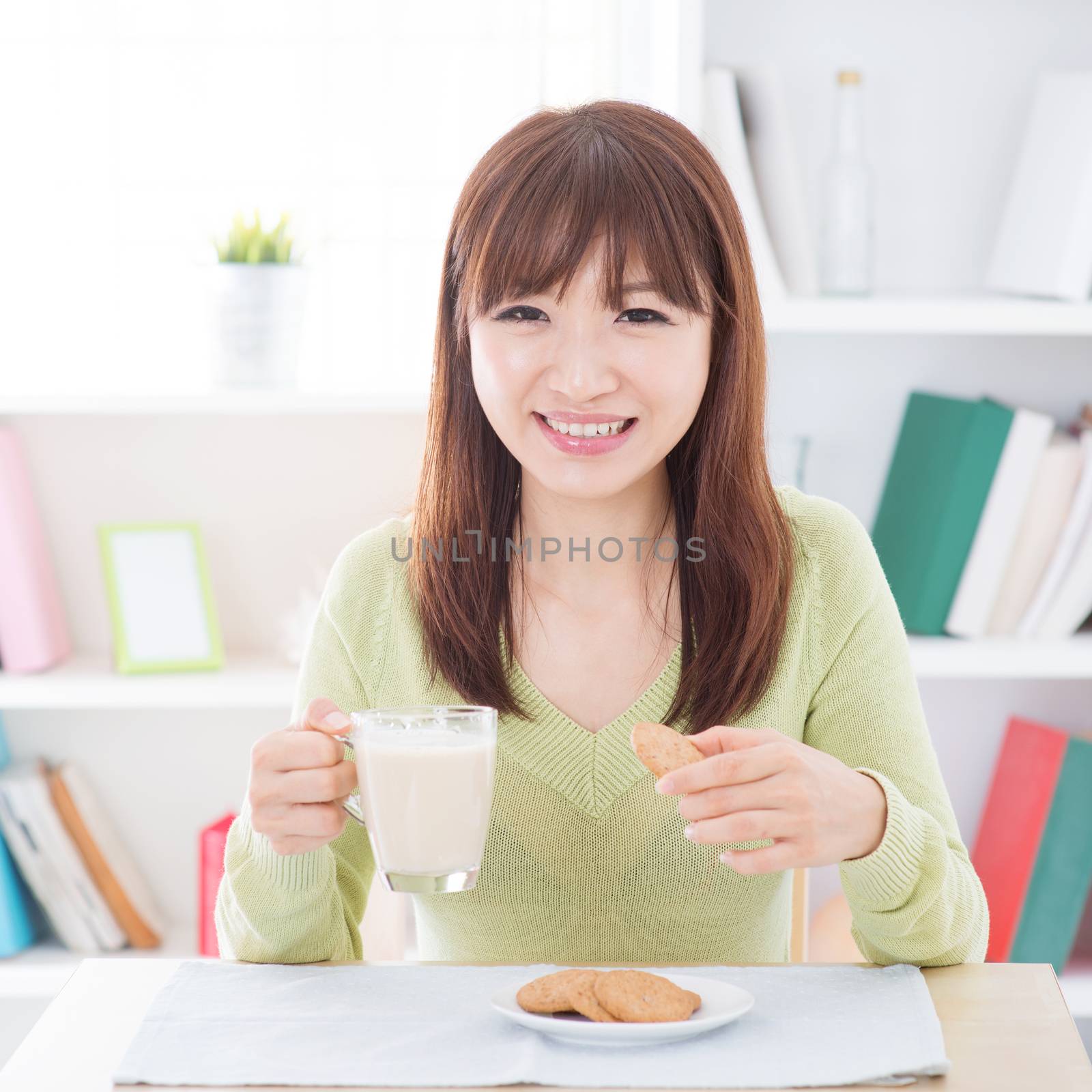 Portrait of happy Asian girl having soymilk and cookies as breakfast. Young woman indoors living lifestyle at home.