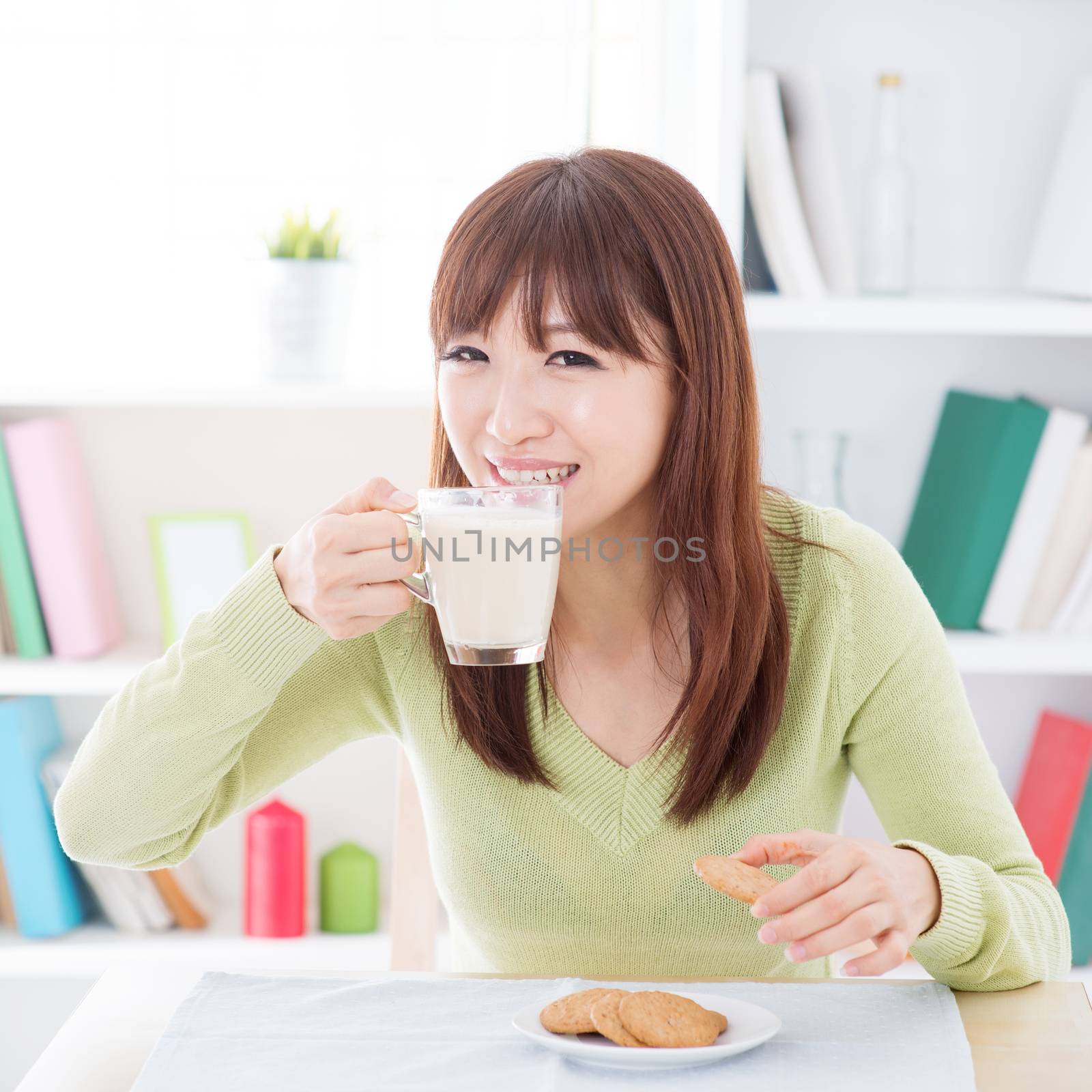 Portrait of happy Asian girl drinking soymilk and having cookies as breakfast. Young woman indoors living lifestyle at home.