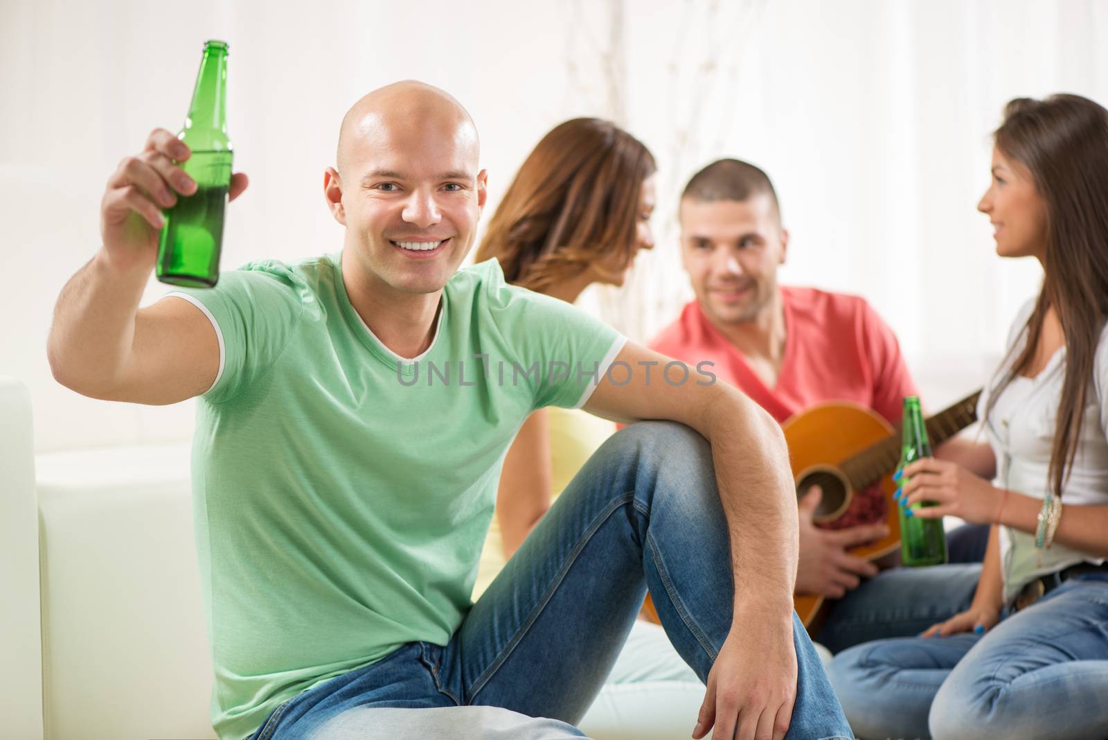 Close up of a young men smiling at home party with beer and cheers. Her friends in the background.