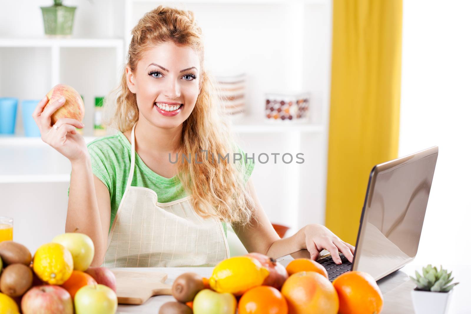 Beautiful young woman holding apple and reading recipe on laptop in kitchen. Looking at camera.