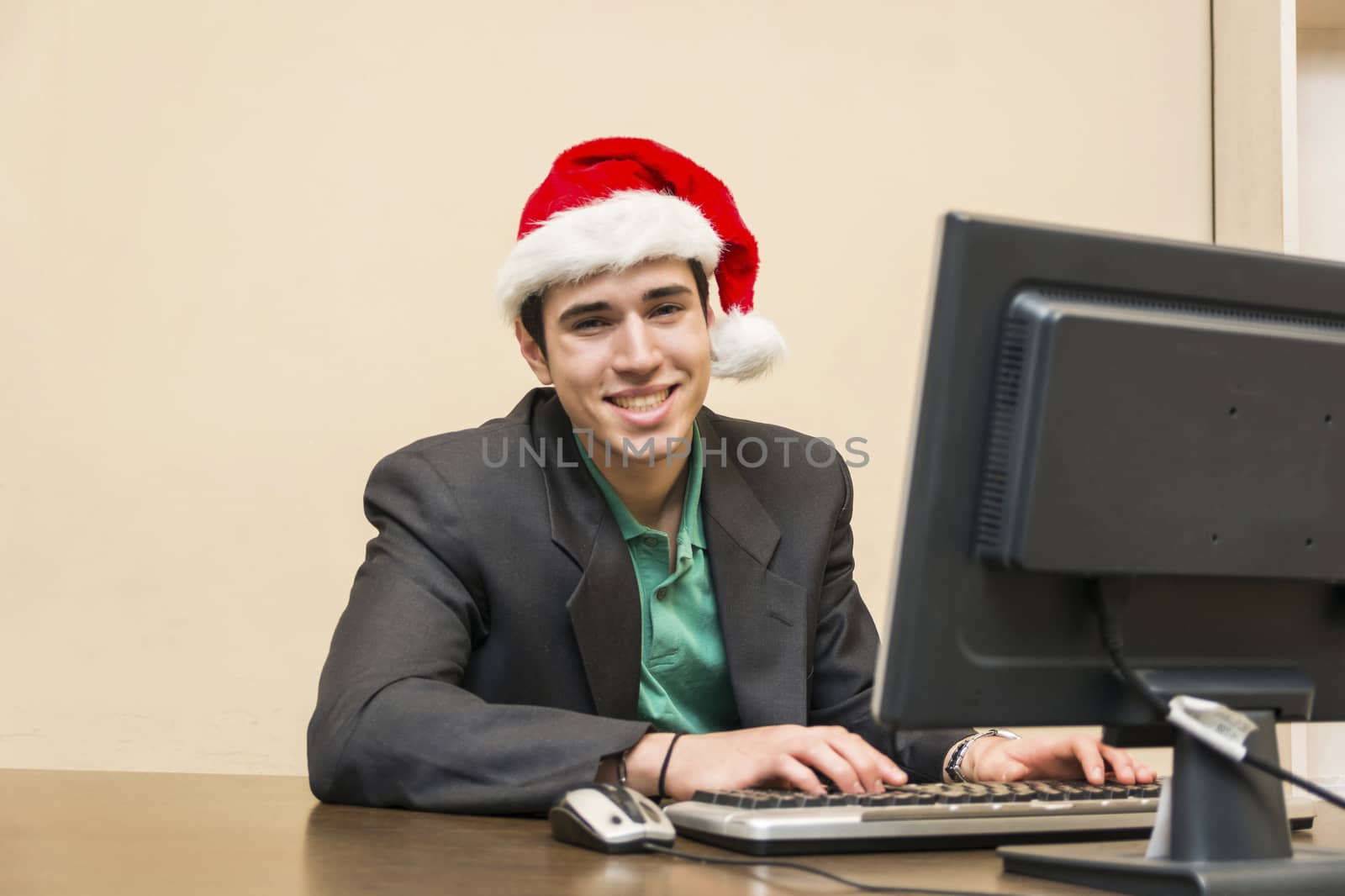 Smiling young businessman sitting at his desk wearing Santa Claus red hat using computer