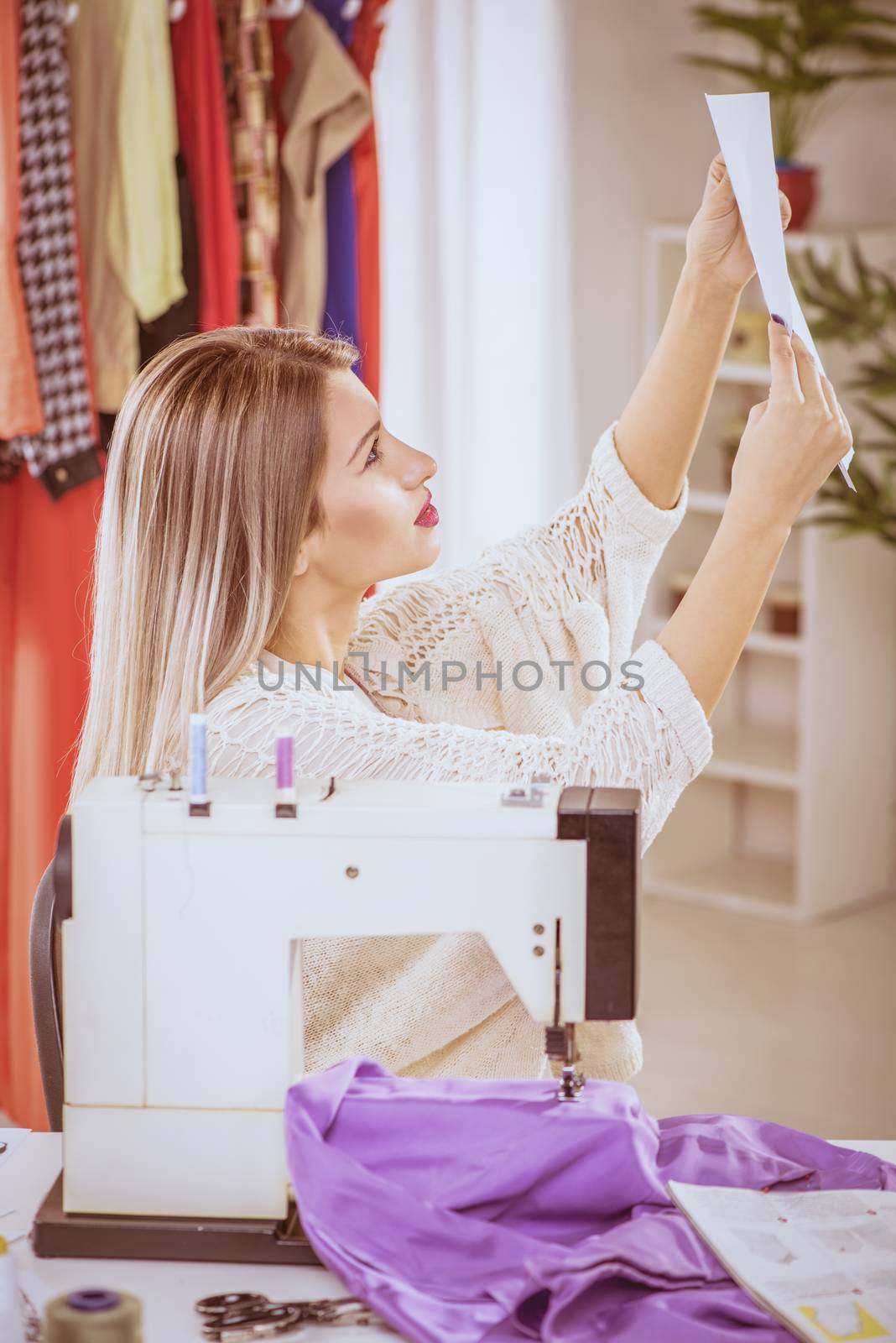 Young woman sits in front of the sewing machine and looking at sketches of clothing.