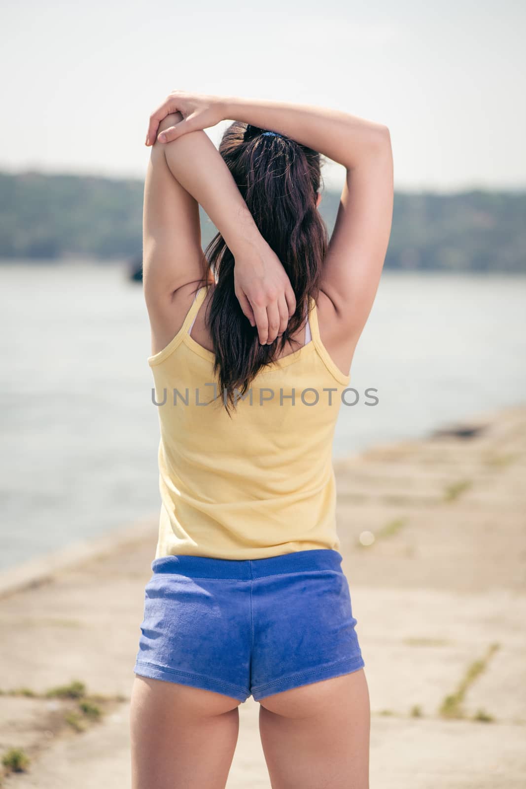 Beautiful young woman doing stretching exercise by the river. Rear view.