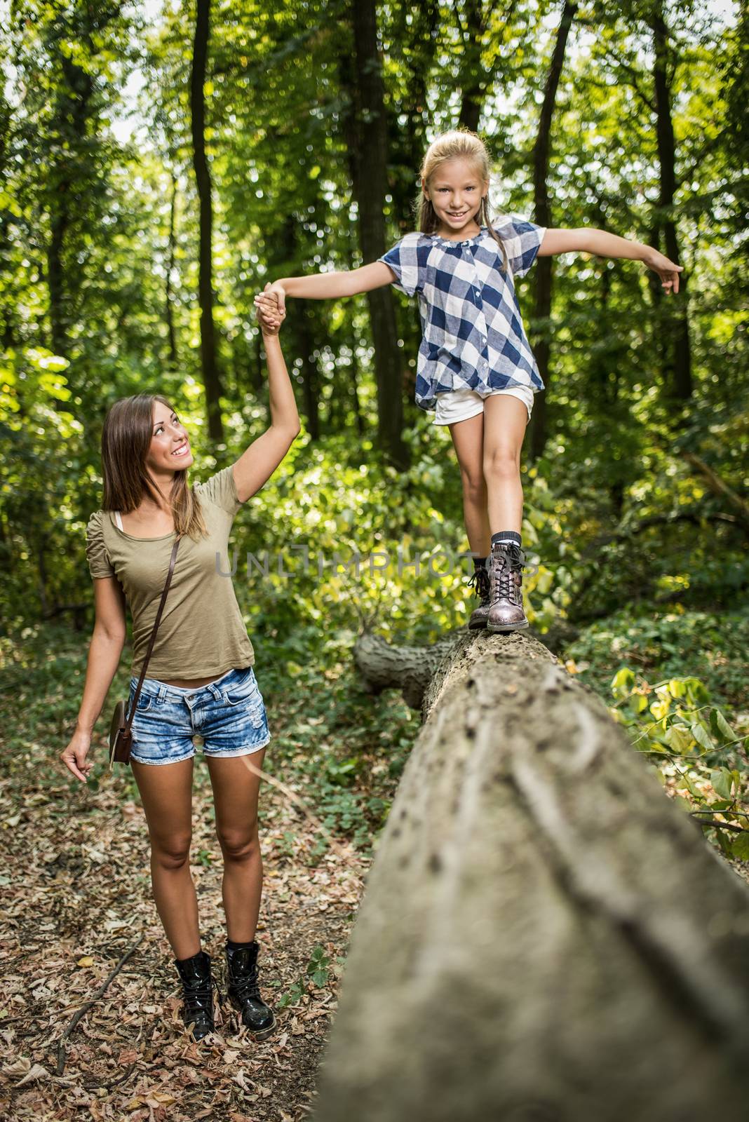 Beautiful young mother and her daughter having fun in the forest. Mother holding girl who walking over fallen tree.
