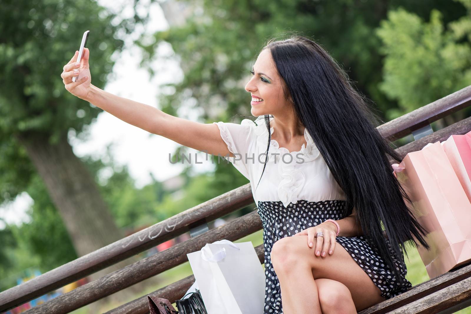 Cheerful girl after shopping with many shopping bags sitting on bench in the park and taking selfie with smart phone.