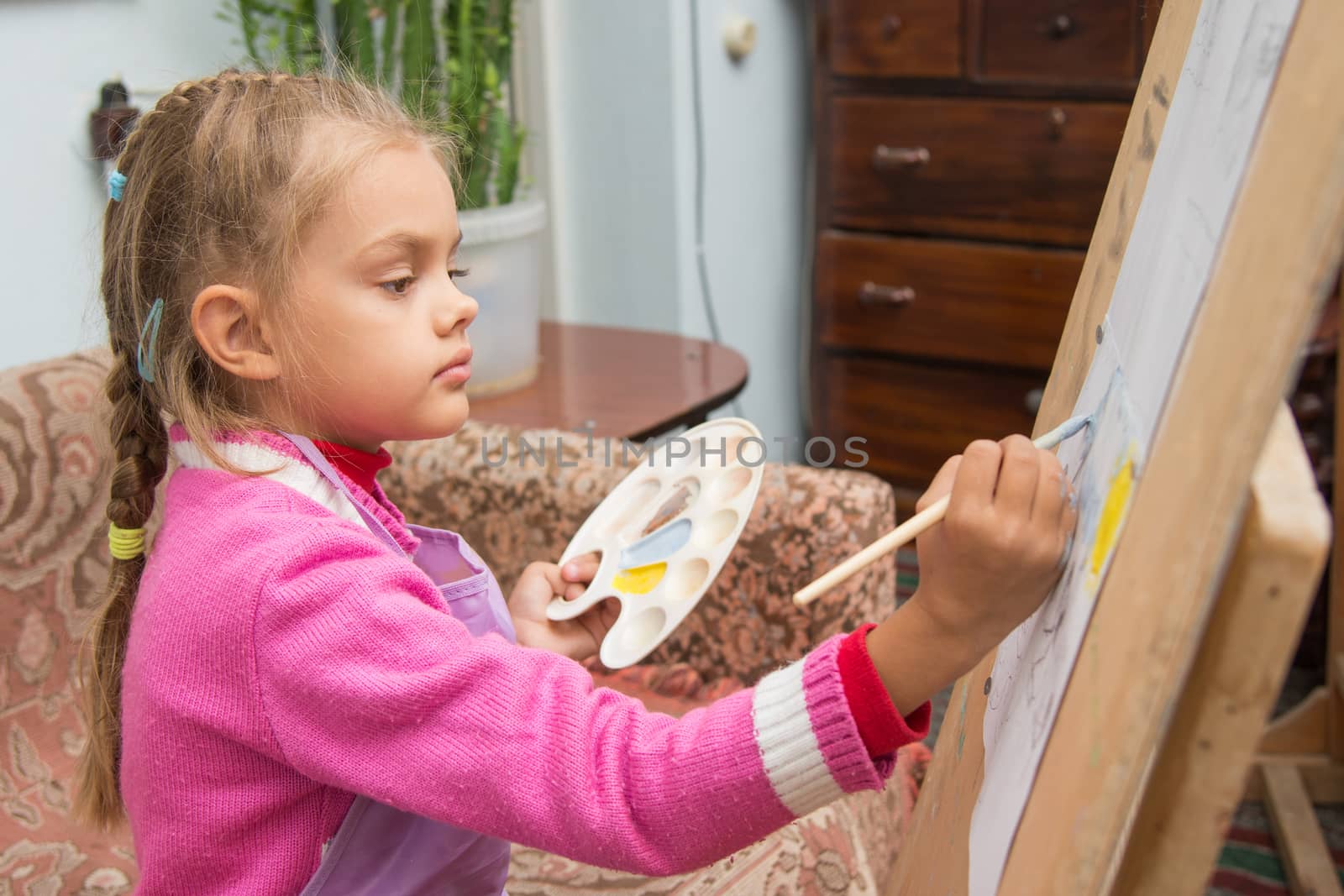 Five-year girl with a brush paints a picture on an easel and holding in his other hand in the artist's palette