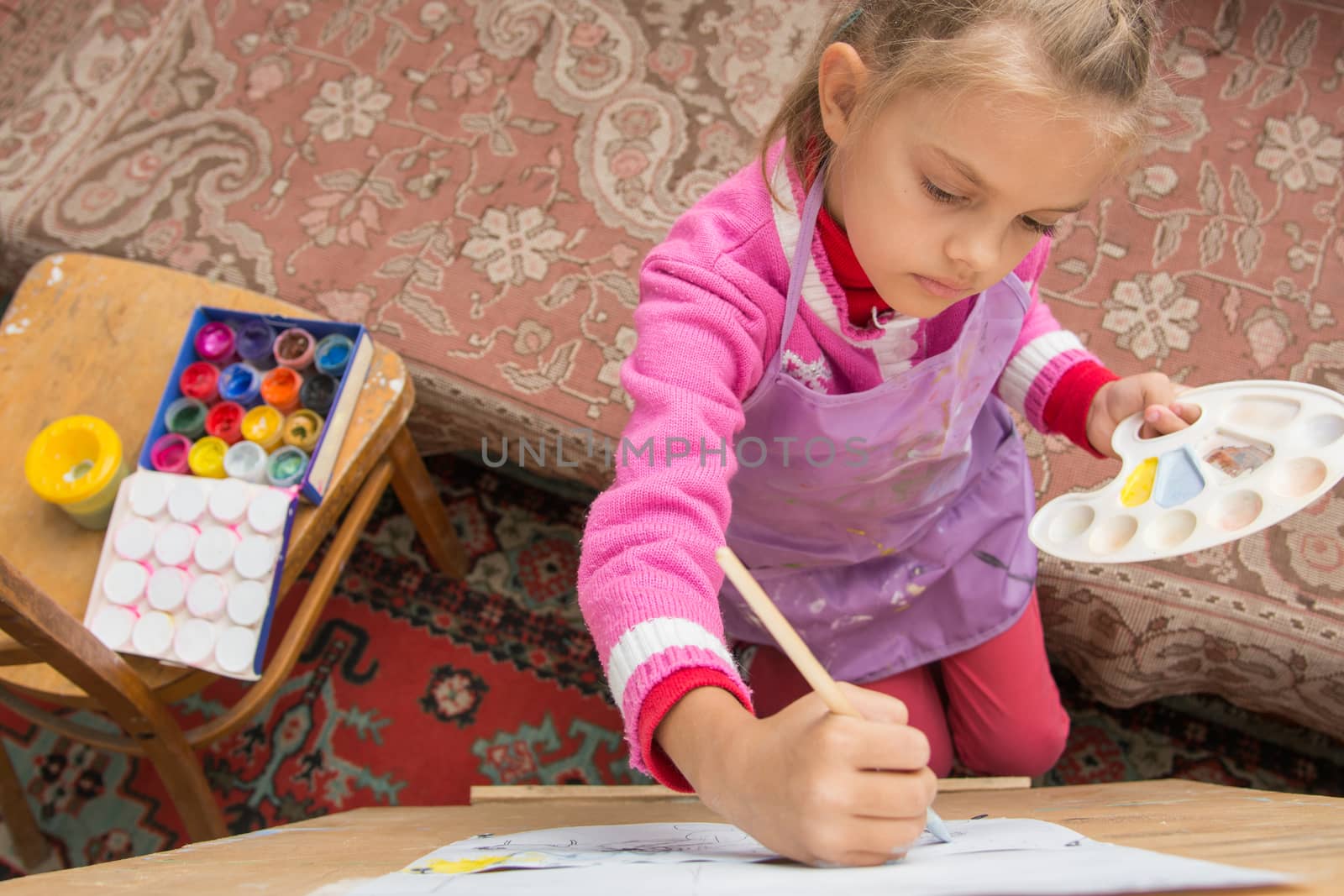 Five-year girl with a brush paints a picture on an easel and holding in his other hand in the artist's palette