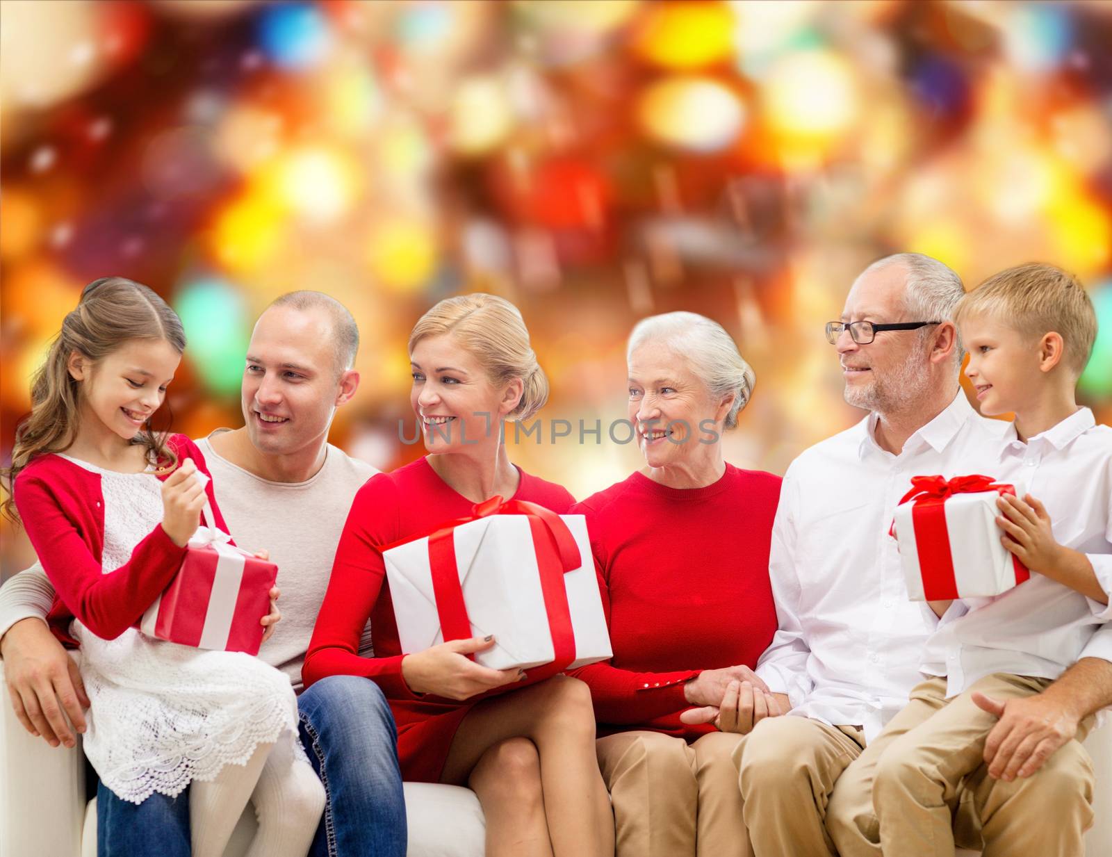 family, holidays, generation, christmas and people concept - smiling family with gift boxes sitting on couch over red lights background