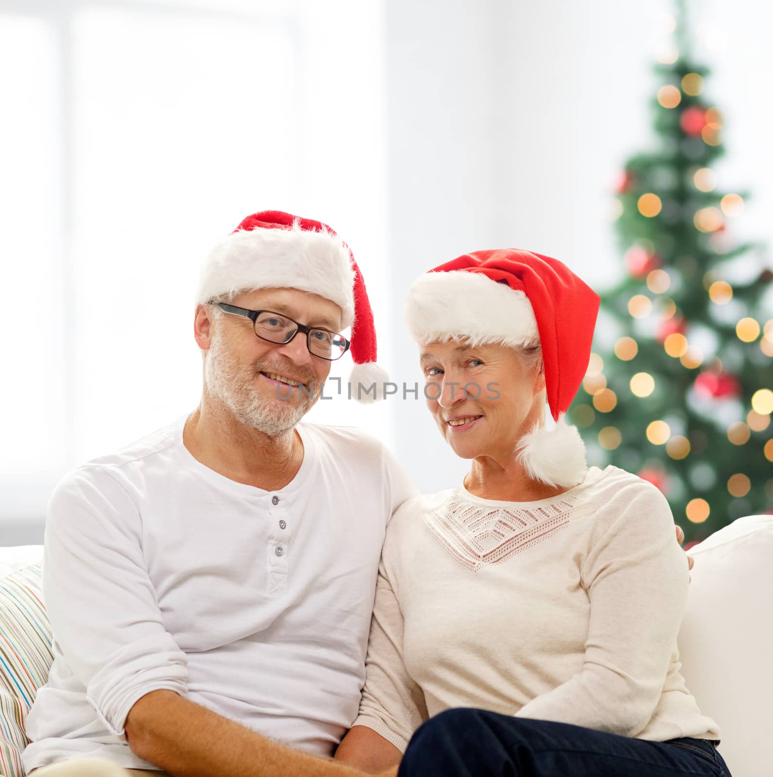 family, holidays, christmas, age and people concept - happy senior couple in santa helper hats sitting on sofa over living room and christmas tree background