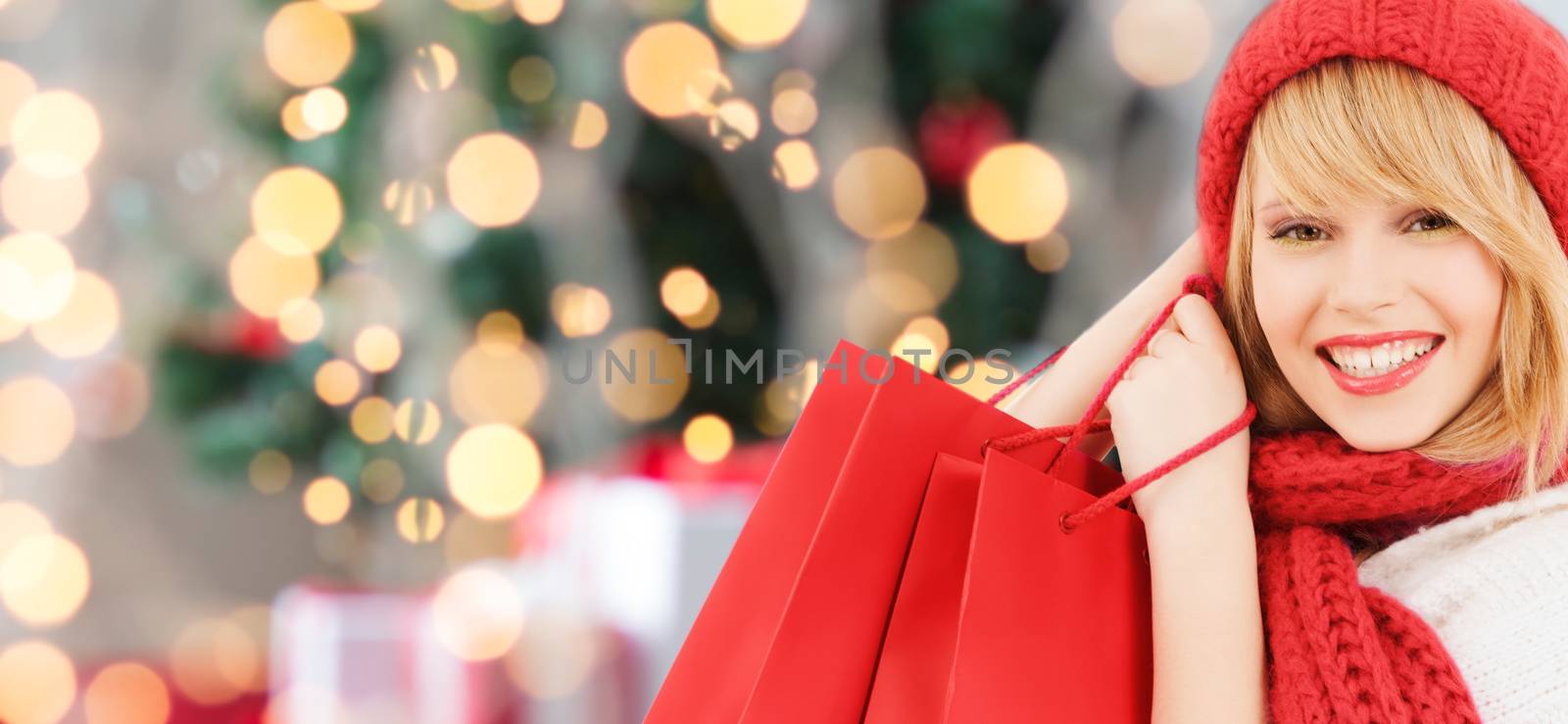 happiness, winter holidays and people concept - smiling young woman in hat and scarf with red shopping bags over christmas tree background