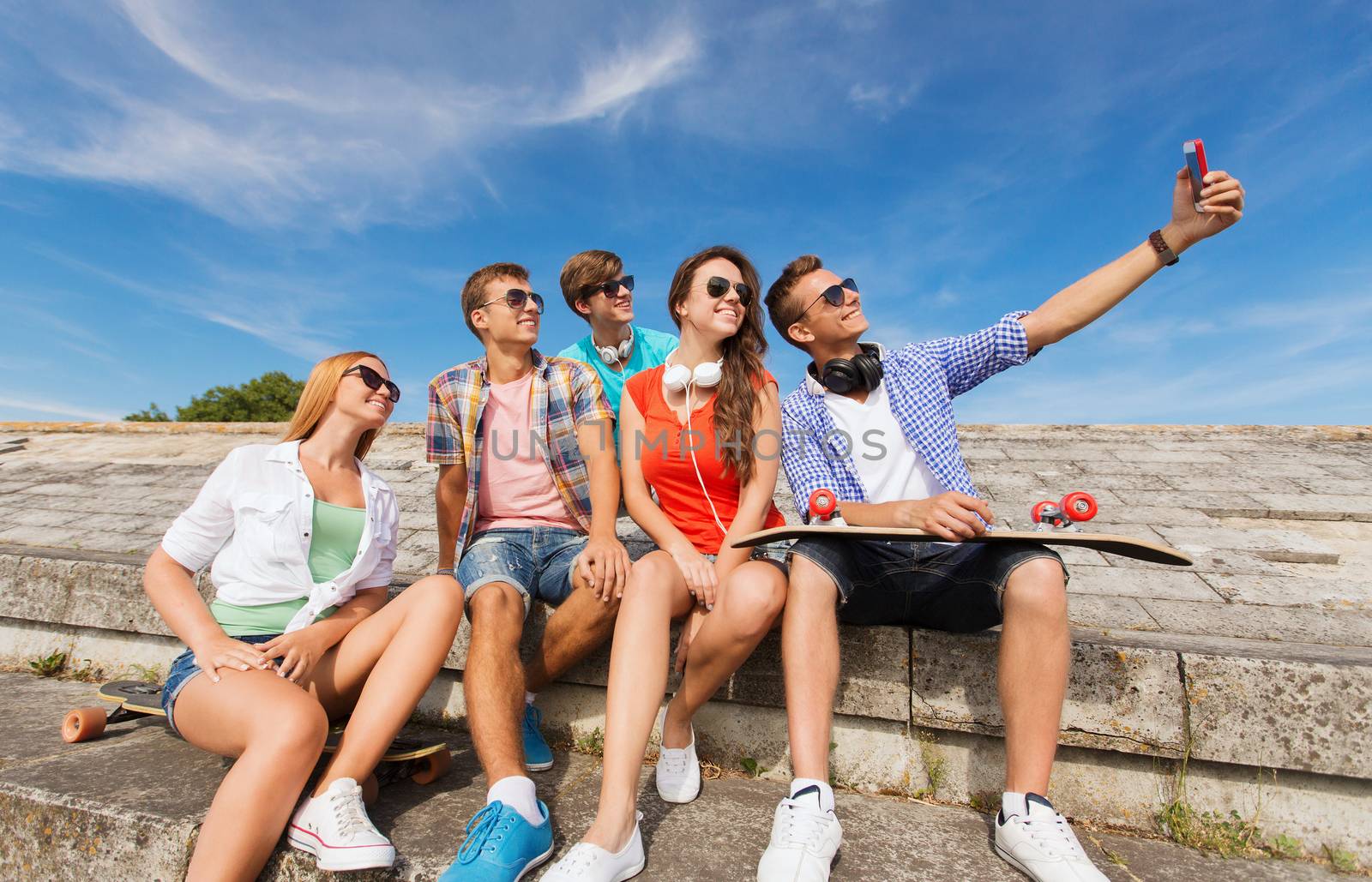 friendship, leisure, summer, technology and people concept - group of smiling friends with skateboard and smartphone making selfie outdoors
