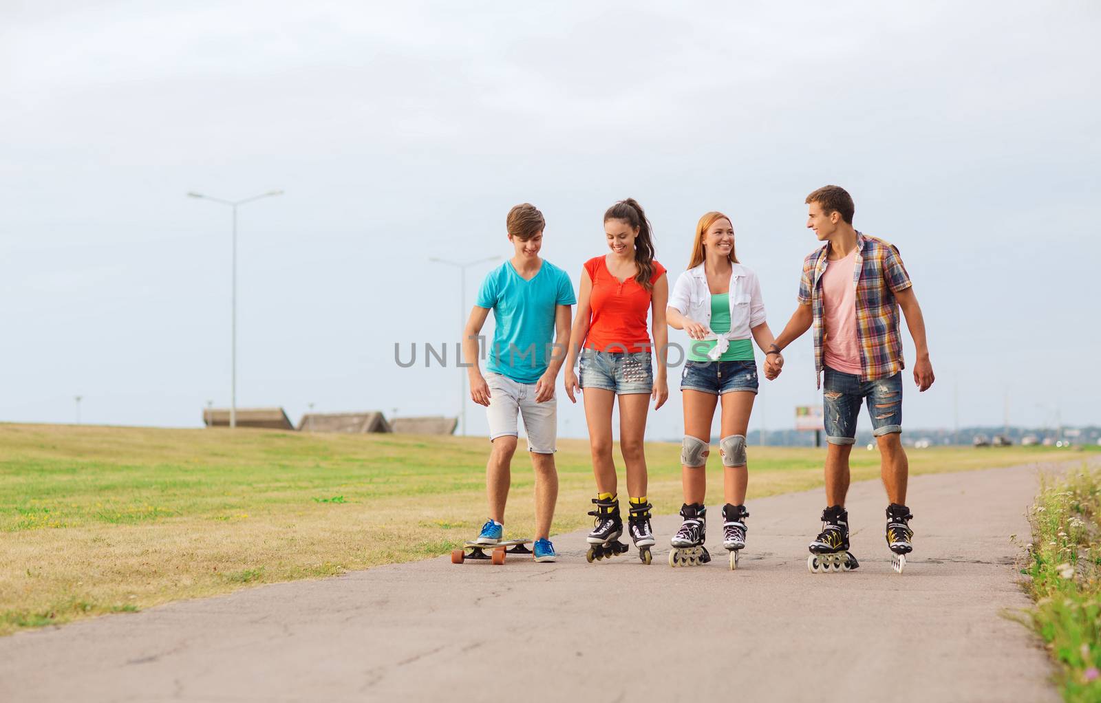 holidays, vacation, love and friendship concept - group of smiling teenagers with roller skates and skateboard riding outdoors