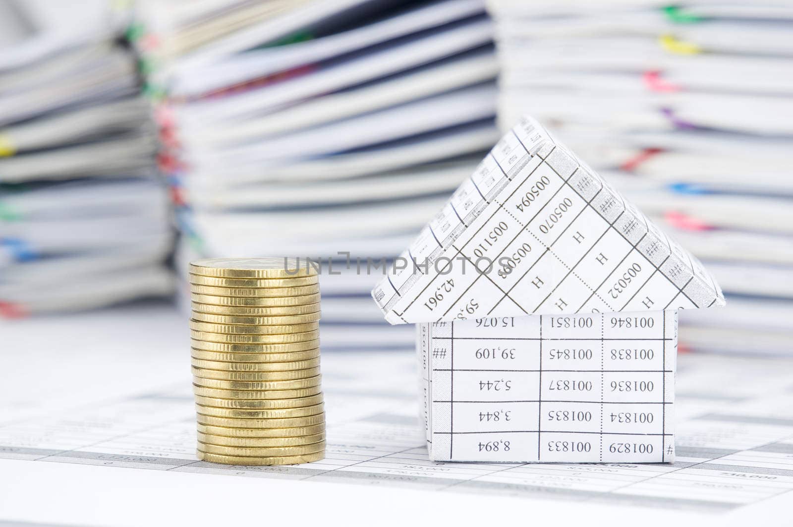 Pile of gold coins and house on finance account have blur pile of paperwork as background.