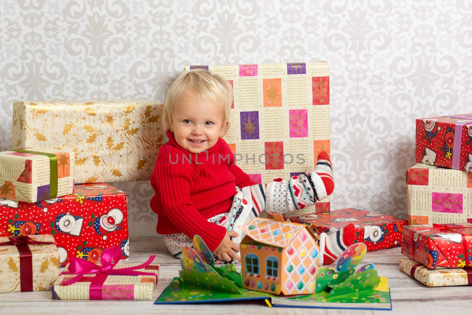 Dressed festively girl with stacks of present boxes around sitting on the floor and smiling.