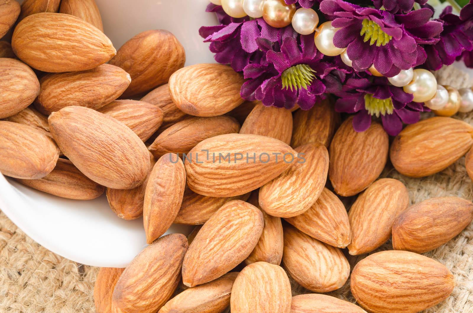 Raw almonds spilling out of small white bowl with flower on sack background.